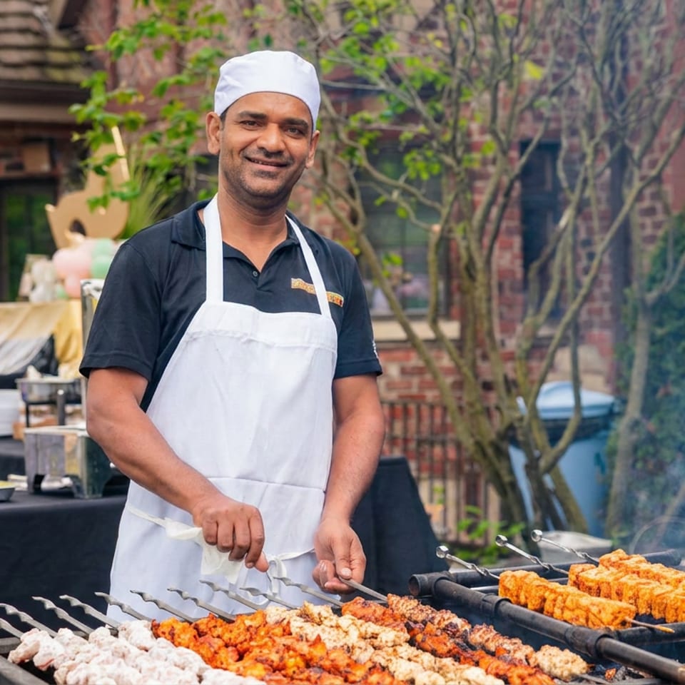 Smiling chef in a white apron grills colorful skewers outdoors while another cook works nearby.