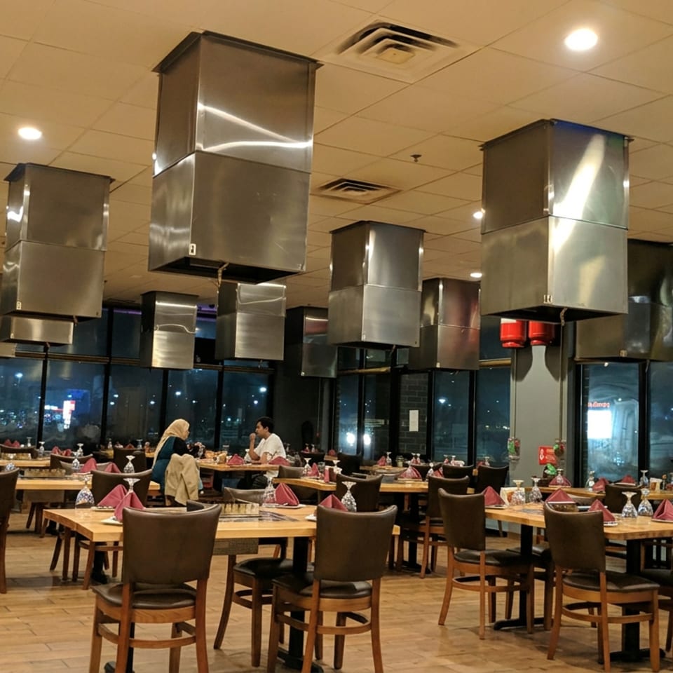 Spacious restaurant dining room with wooden tables, folded napkins, seated diners, and numerous overhead stainless-steel ventilation hoods.