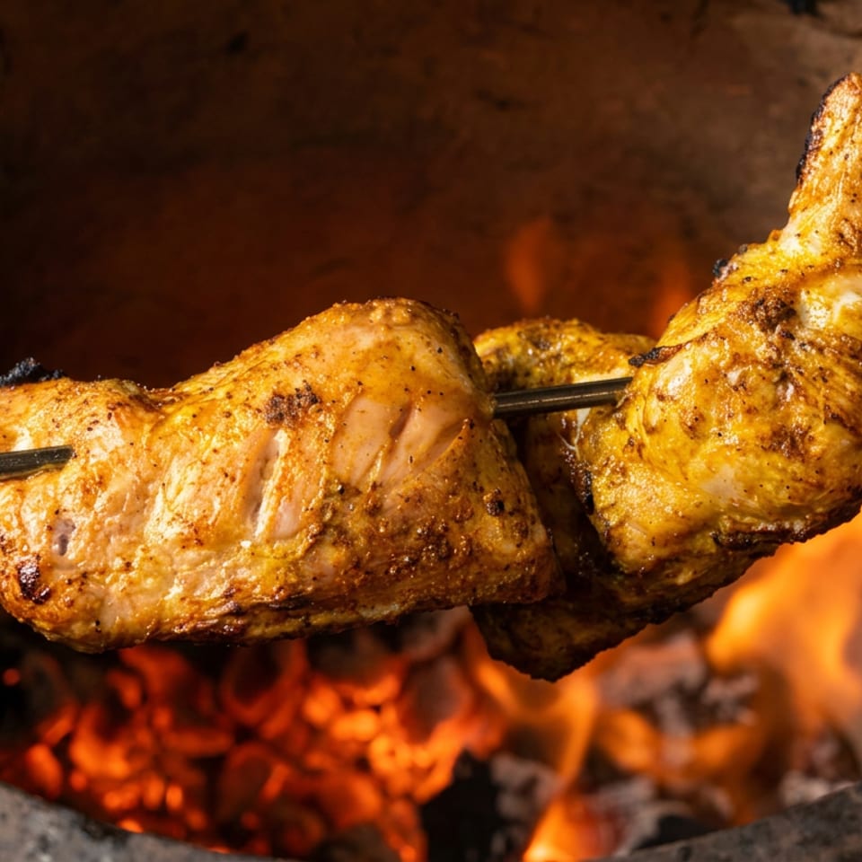 Marinated chicken pieces roast on a skewer over glowing charcoal and open flames in a tandoor oven.