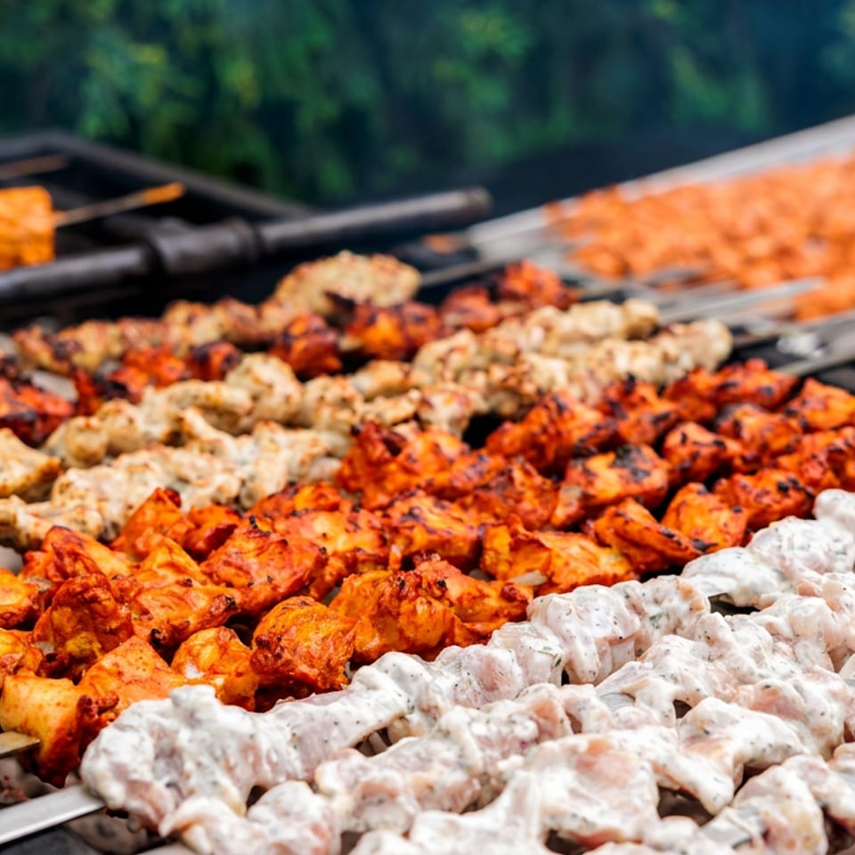 Rows of colorful marinated meat skewers grilling over charcoal outdoors, including orange, red, beige, and white varieties.