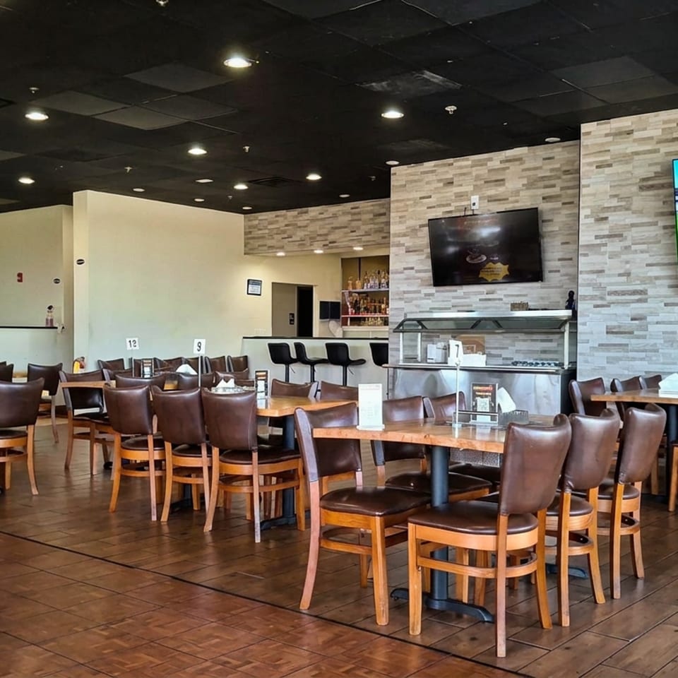 Spacious restaurant dining room with wood tables, leather chairs, bar counter, buffet display, and televisions showing a sporting event.