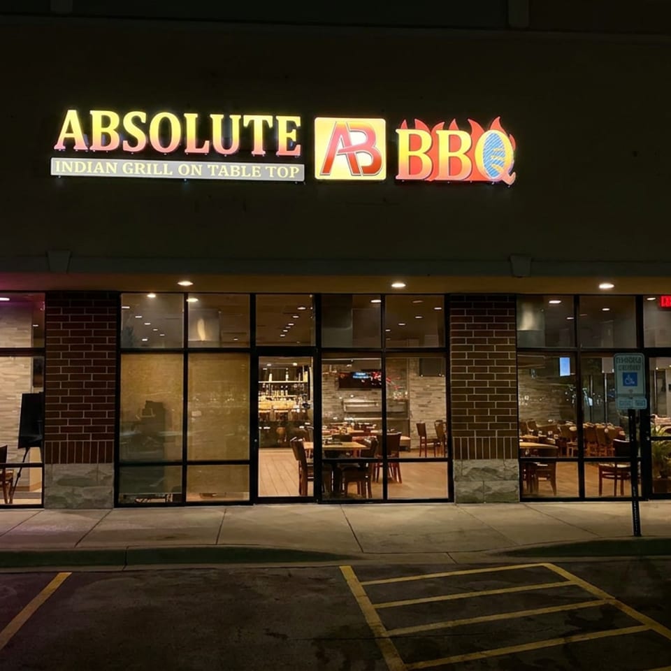 Nighttime exterior of Absolute BarBQ, an Indian grill restaurant, with illuminated signage, glass windows, and visible dining tables.