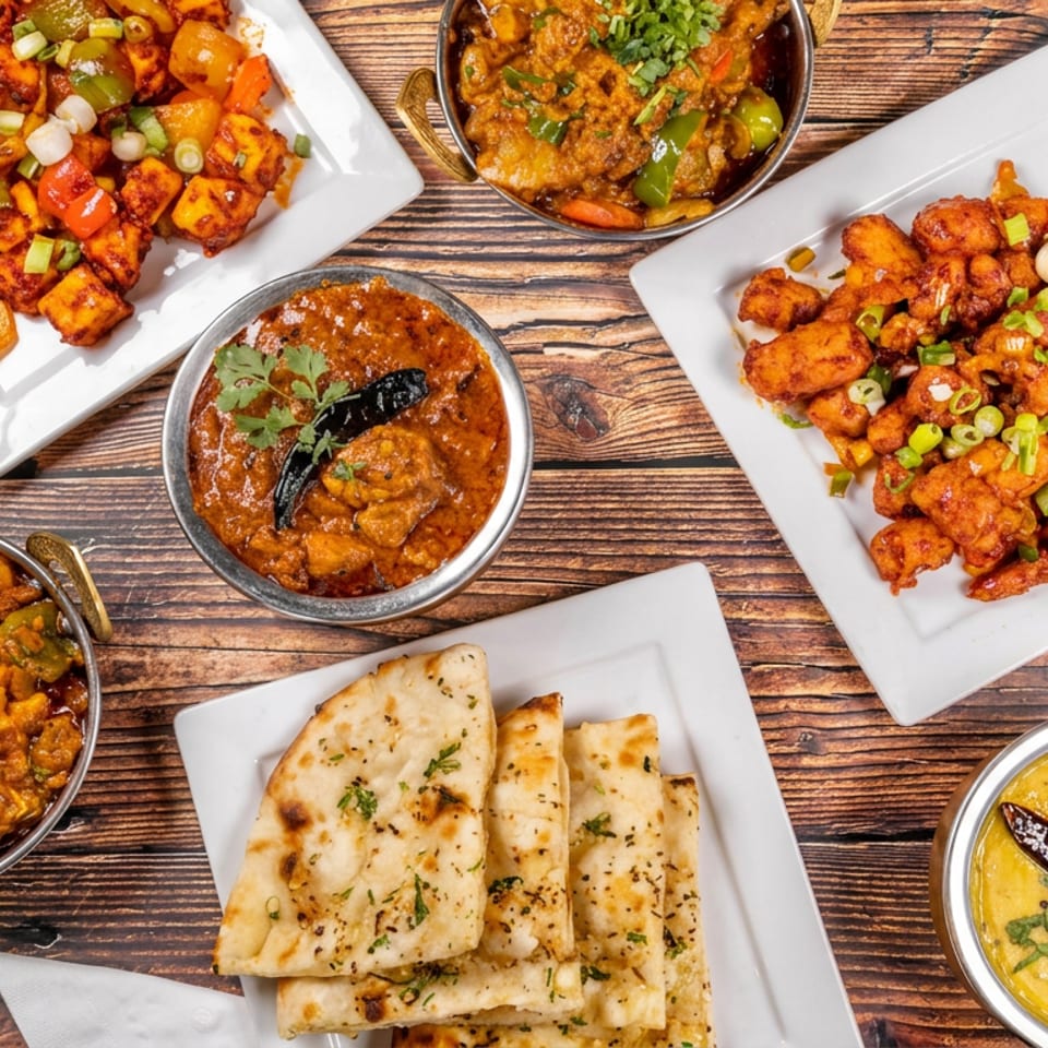 Overhead view of an Indian meal spread with curries, fried appetizers, garnished vegetables, and naan arranged on a wooden table.