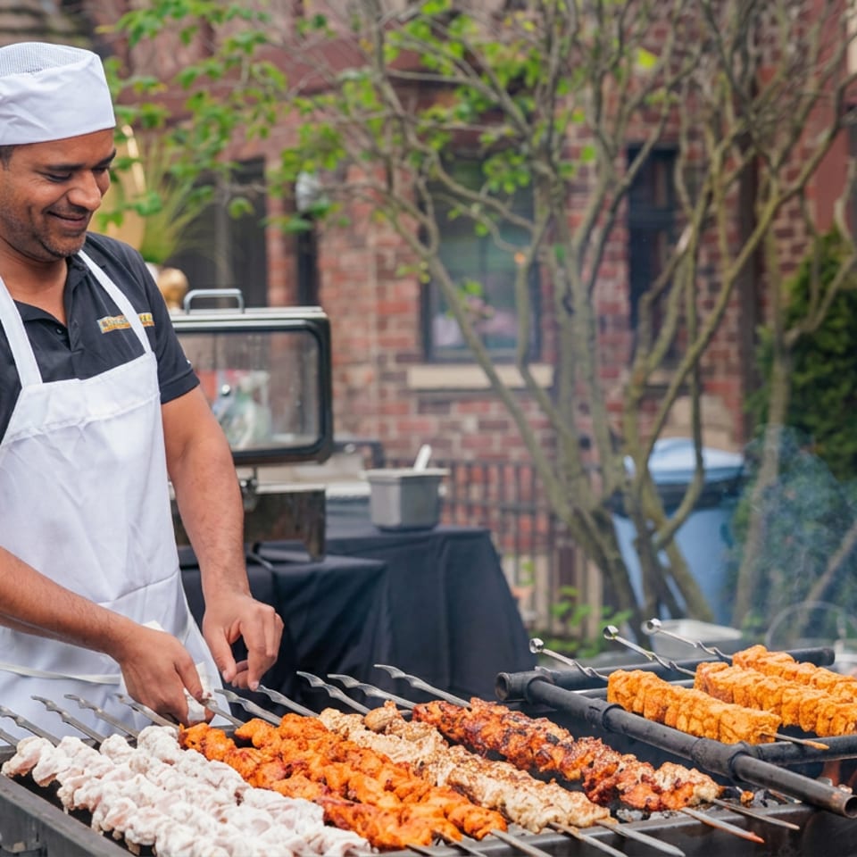 Chef grilling colorful skewered meats at an outdoor restaurant barbecue while guests gather in a leafy courtyard.