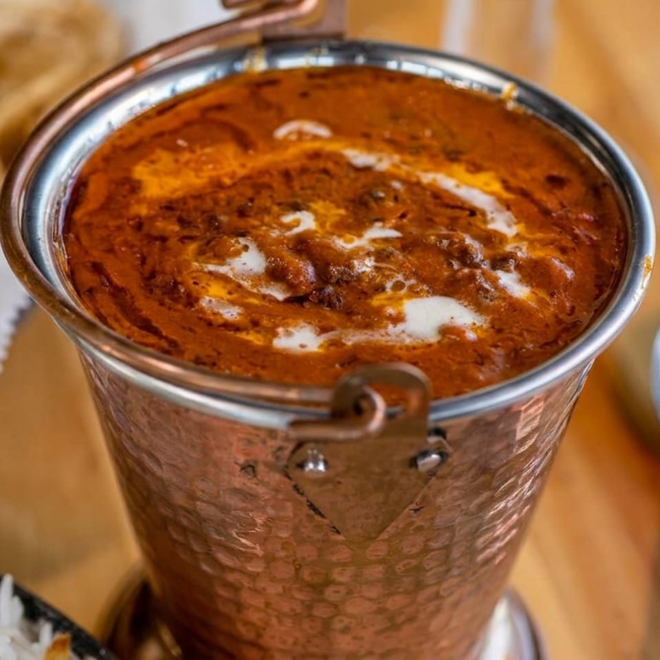 Copper bowl of creamy orange curry garnished with cream, served with naan, rice, and a spoon.
