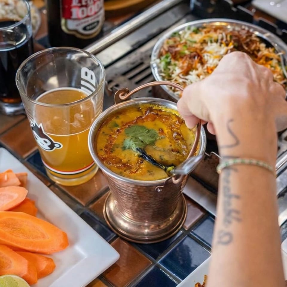 Hand serves creamy soup from a metal pot beside sliced sweet potatoes, drinks, and a tabletop grill.